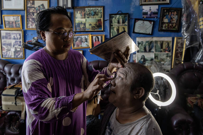 A imagem mostra um artista de maquiagem aplicando produtos no rosto de uma pessoa em um estúdio. O artista está usando uma camisa roxa e está concentrado no trabalho. O fundo é decorado com várias fotos emolduradas e há uma luz circular iluminando a cena.