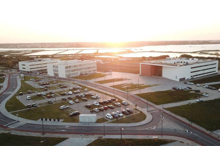 A imagem mostra uma vista aérea de um complexo de edifícios modernos, com um grande estacionamento em primeiro plano. O sol está se pondo ao fundo, iluminando a cena. Há várias áreas verdes ao redor e um corpo d'água visível ao fundo, com campos e casas ao longe. O estacionamento está cheio de carros e há ruas bem definidas ao redor dos edifícios.