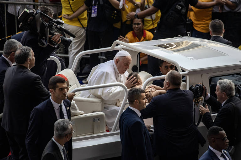 A imagem mostra o Papa sentado em um carro aberto, cercado por segurança e pessoas ao seu redor. Ele está em um evento público, com uma multidão visível ao fundo, incluindo pessoas vestindo camisetas amarelas. O ambiente parece ser festivo e animado