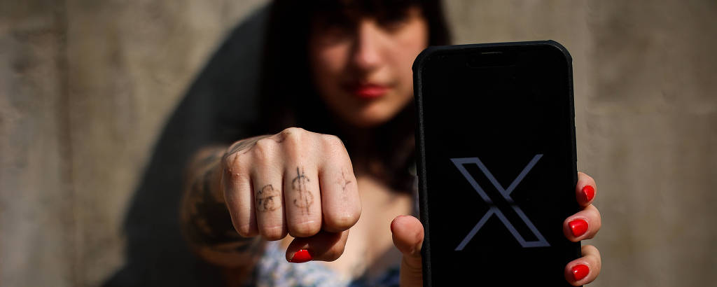 Uma pessoa está segurando um celular com a tela voltada para a câmera, exibindo o logo 'X'. A pessoa tem cabelo escuro e liso, e está usando uma blusa de alças. Ela está com a mão esquerda fechada em um punho, mostrando tatuagens. O fundo é uma parede de concreto.