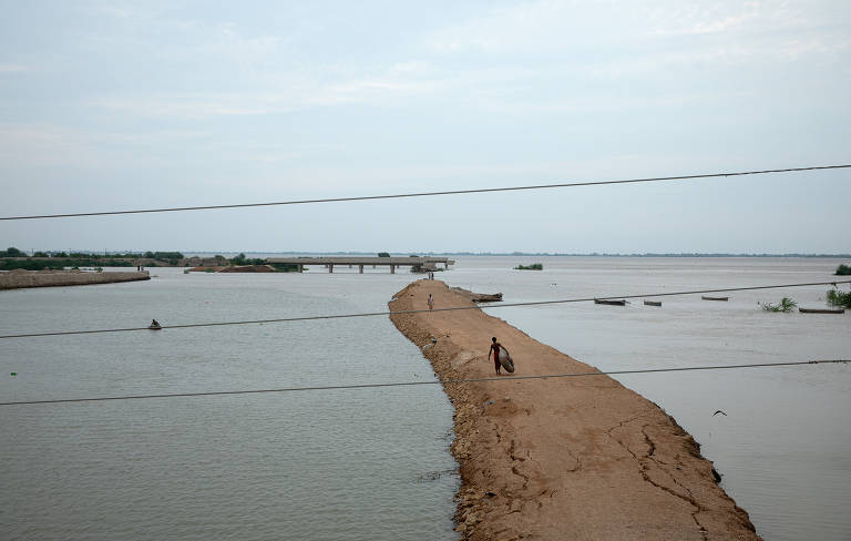 A imagem mostra um caminho de terra estreito que se estende por uma área alagada. À esquerda, há uma ponte visível ao fundo, enquanto pequenas embarcações estão flutuando na água. O céu está nublado, e uma pessoa está caminhando ao longo do caminho de terra. A paisagem é predominantemente aquática, com vegetação ao redor.
