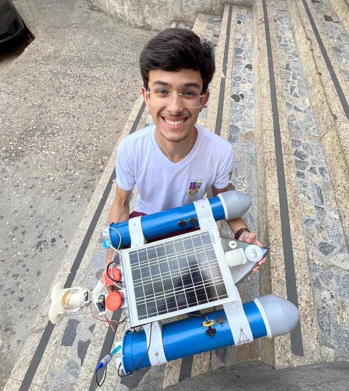 jovem de óculos sorri para a foto, segurando aparelho com placa solar e canos de PVC de cor azul