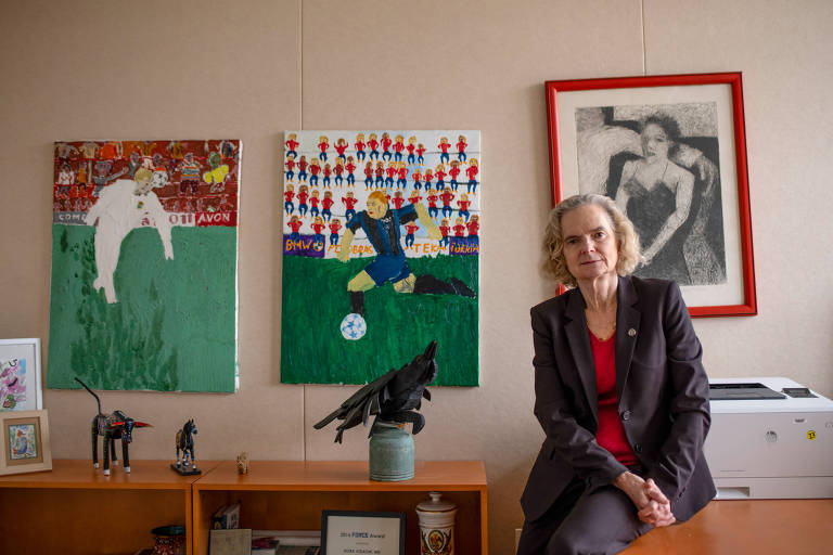 Uma mulher sentada em um escritório, vestindo um terno escuro e uma blusa vermelha. Ao fundo, há três obras de arte: duas pinturas coloridas relacionadas ao futebol e uma imagem em preto e branco de uma mulher. A mesa ao lado contém uma impressora e alguns objetos decorativos.
