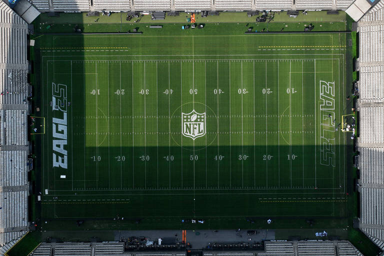 Imagem aérea de um campo de futebol americano, com grama verde bem cuidada. As linhas do campo estão claramente marcadas, e há o logotipo 'EAGL' em uma das extremidades. O estádio ao redor é visível, mas vazio.
