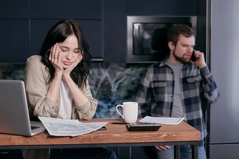 A imagem mostra uma mulher sentada à mesa com expressão preocupada, apoiando o rosto nas mãos. Ela está cercada por papéis e um laptop aberto. Ao fundo, um homem está de pé, falando ao telefone, com uma expressão séria. Uma caneca branca e uma calculadora estão sobre a mesa, junto a mais papéis. O ambiente é uma cozinha moderna com armários escuros.