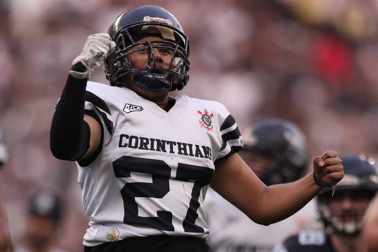 Um jogador de futebol americano, vestindo um uniforme branco e preto com a palavra 'CORINTHIAN' e o número 27, está comemorando com um punho cerrado. Ele usa um capacete e parece estar em um campo de jogo, com outros jogadores ao fundo. A atmosfera é de celebração e energia.
