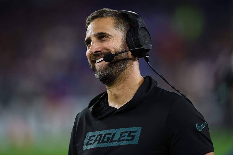 Um homem sorridente, usando um fone de ouvido e uma camiseta preta com a palavra 'EAGLES' estampada. Ele parece estar em um ambiente de jogo, possivelmente em um estádio, com um fundo desfocado que sugere uma partida de futebol americano.
