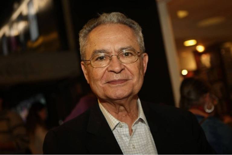 Um homem idoso com cabelo grisalho e óculos, vestindo uma camisa clara e um paletó escuro. Ele está sorrindo levemente e olhando para a câmera. Ao fundo, há pessoas e uma iluminação suave, sugerindo um ambiente social ou de evento.