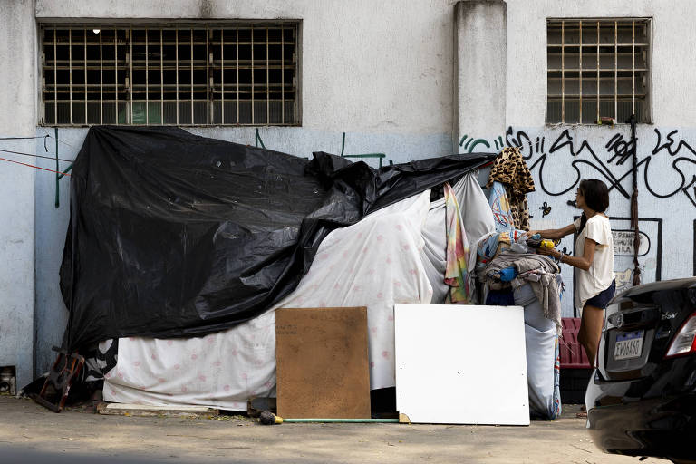 A imagem mostra um abrigo improvisado feito com lonas pretas e cobertores, localizado em uma área urbana. Ao lado do abrigo, uma pessoa está organizando objetos. Ao fundo, há uma parede com janelas gradeadas e grafites. No chão, há painéis de madeira e um carro estacionado