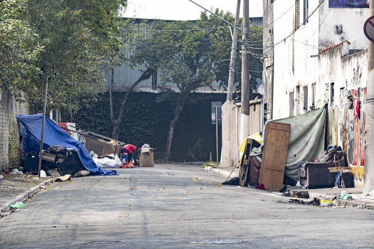 A imagem mostra uma rua estreita e deserta, com barracas feitas de lonas azuis e verdes ao lado da calçada. No fundo, uma pessoa está agachada, aparentemente organizando seus pertences. Há móveis e objetos espalhados pelo chão, incluindo um sofá e caixas. As paredes dos edifícios próximos estão desgastadas e há vegetação ao fundo.