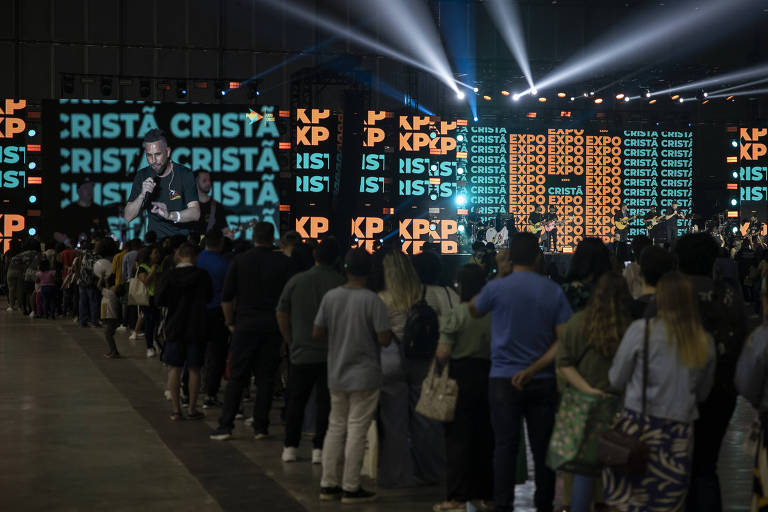 A imagem mostra um evento de música cristã com um palco iluminado e uma grande tela de fundo exibindo as palavras 'EXPO CRISTÃ'. Há uma multidão em fila, assistindo a um artista que está se apresentando no palco. O artista é visto em uma tela grande, enquanto no palco há outros músicos. O ambiente é bem iluminado com luzes coloridas.
