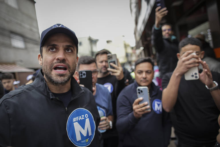 Fotografia mostra o candidato Pablo Marçal, um homem branco com barba preta, em primeiro plano, vestindo uma jaqueta preta e um boné azul marinho, com um emblema azul com a letra 'M'. Ele parece estar falando ou se expressando de forma intensa. Atrás dele, há um grupo de pessoas, algumas segurando celulares, que parecem estar filmando ou tirando fotos. O ambiente é urbano, com edifícios ao fundo.