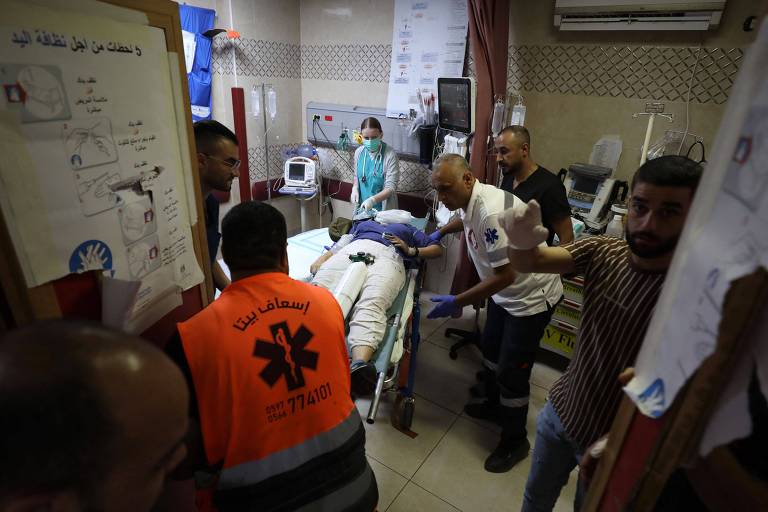 A imagem mostra um ambiente hospitalar onde uma equipe médica está prestando atendimento a um paciente em uma maca. Várias pessoas estão ao redor, algumas vestindo uniformes de emergência. O paciente está deitado de bruços, enquanto os profissionais de saúde parecem estar realizando procedimentos médicos. O ambiente é iluminado e há equipamentos médicos visíveis ao fundo.