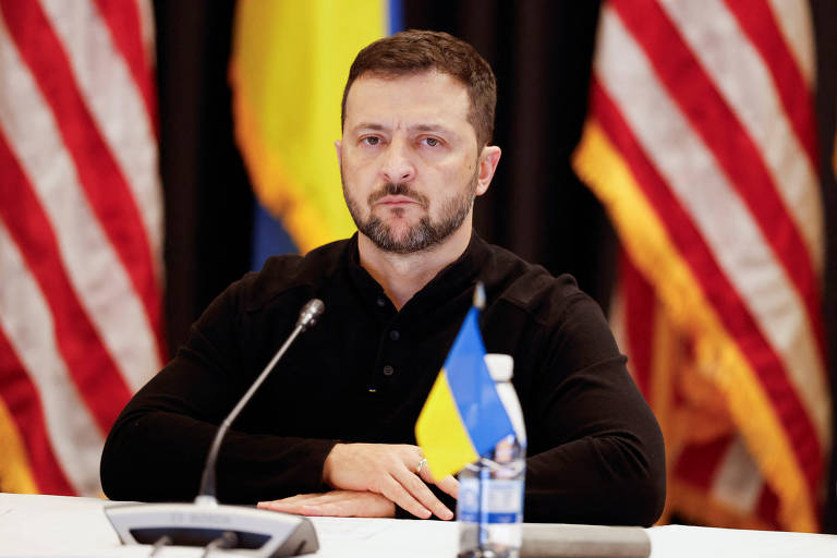 Volodimir Zelenski, homem branco de barba e cabelos escuros, está sentado em frente a uma mesa com uma bandeirinha amarela e azul da Ucrânia, microfone e garrafa d'água. Ele usa uma malha escura. Ao fundo, bandeiras dos EUA e da Ucrânia