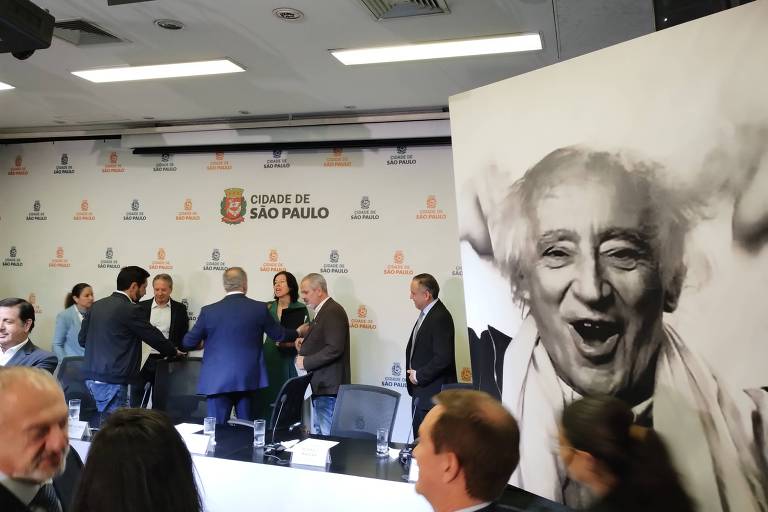 A imagem mostra um evento em um auditório, com várias pessoas interagindo em um painel. Ao fundo, há uma grande foto em preto e branco de um homem sorridente. O ambiente é formal, com mesas e cadeiras dispostas para os participantes. O logotipo de uma instituição está visível na parede ao fundo.