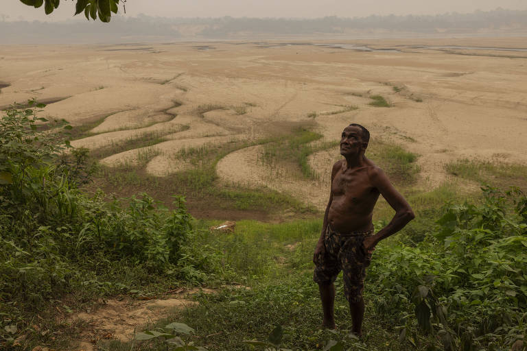 Homem sem camisa em barranco com pouca vegetação e, ao fundo, o rio madeira com imensos bancos de areia; quase não há água; o céu está cheio de fumaça