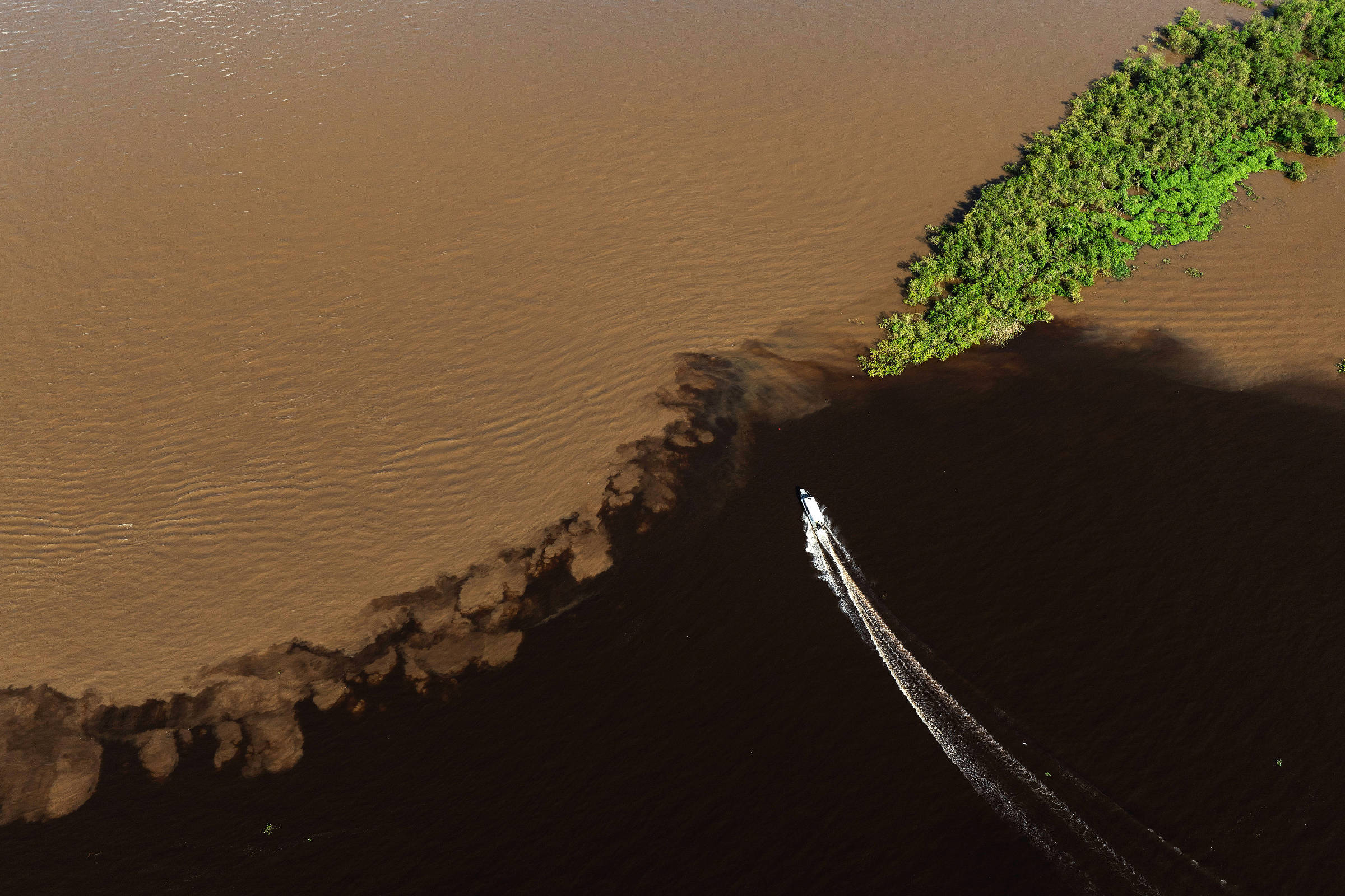 Barco navega em água escura ao lado de porção com água marrom e barrenta com vegetação flutuante