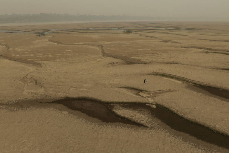 A imagem mostra uma vasta área de areia seca com padrões de ranhuras e pequenas depressões. No centro, há uma figura humana pequena caminhando sobre a superfície. O céu está cinza, e a atmosfera cheia de fumaça. Ao fundo, é possível ver uma linha de água
