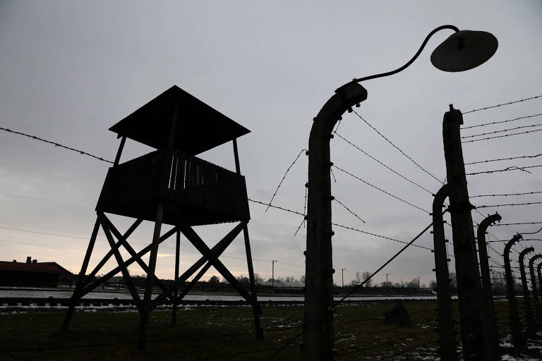 A imagem mostra uma torre de vigilância escura, situada em um campo de concentração, com cercas de arame farpado ao redor. O céu está nublado, criando uma atmosfera sombria. Uma lâmpada de poste está visível ao lado da torre