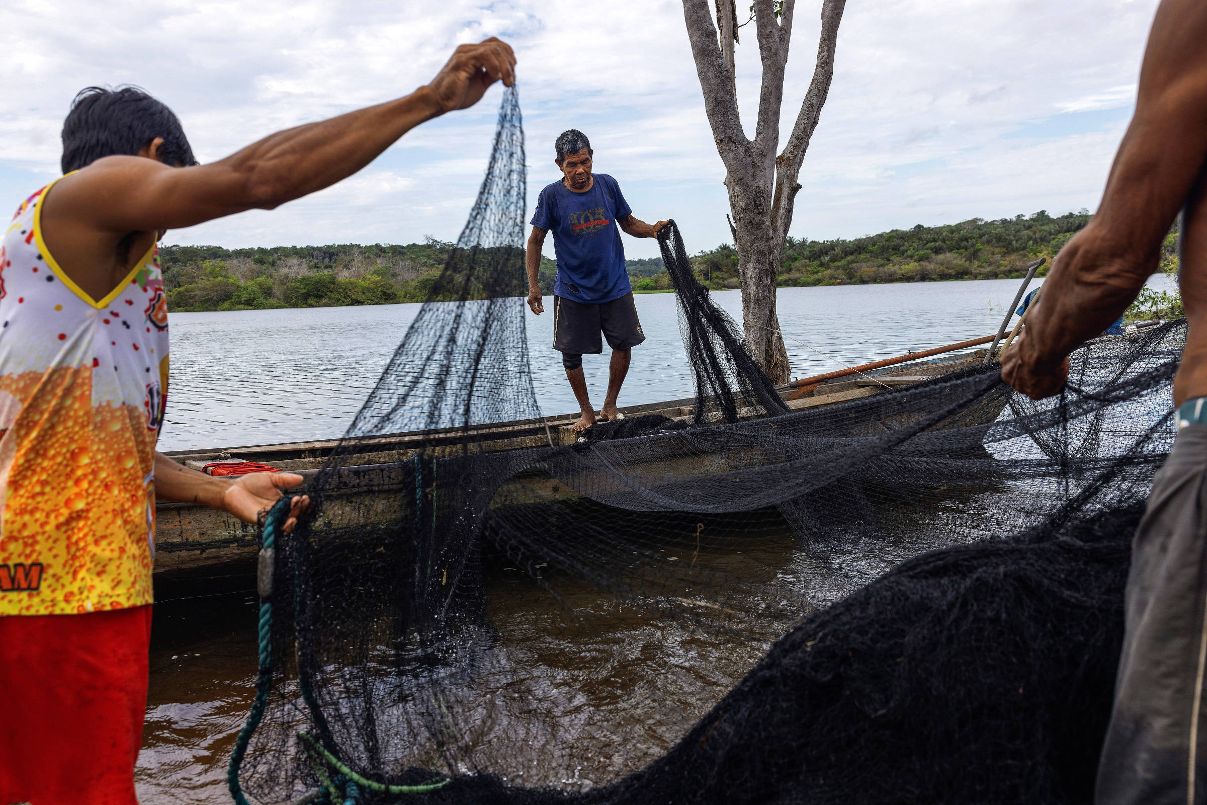 Três homens seguram redes de pesca na beira de um rio