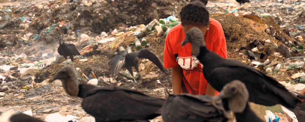 A imagem mostra uma criança agachada em um lixão, cercada por urubus. O ambiente é de lixo e resíduos, com a criança vestindo uma camiseta vermelha. Os urubus estão próximos, alguns se alimentando do lixo ao redor. O fundo é uma área desordenada com montanhas de lixo e outros urubus visíveis.