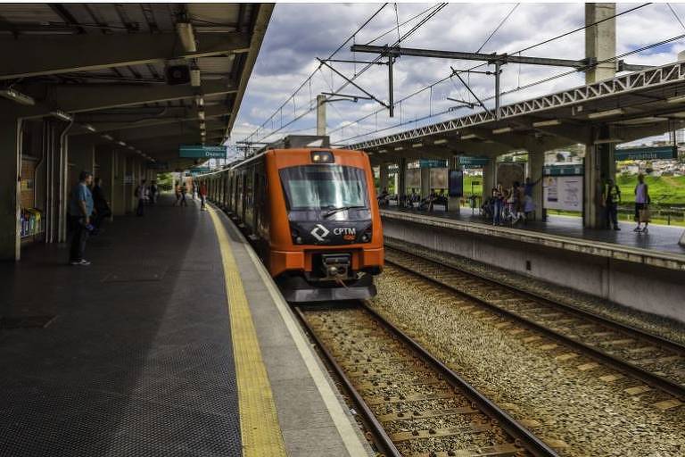 A imagem mostra um trem laranja se aproximando de uma plataforma em uma estação ferroviária. A plataforma é coberta e há pessoas esperando. O céu está nublado, e os trilhos do trem são visíveis ao longo da imagem