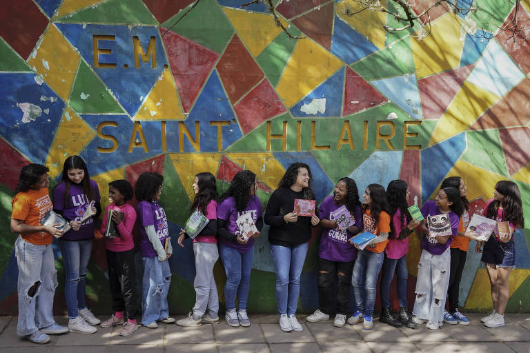 Um grupo de jovens está posando em frente a um mural colorido. Elas estão alinhados, segurando livros e cadernos. As meninas vestem camisetas de diferentes cores, como roxo, laranja e preto. O mural ao fundo apresenta formas geométricas em várias cores, e a frase 'E.M. Saint-Hilaire' está escrita em letras grandes e amarelas.