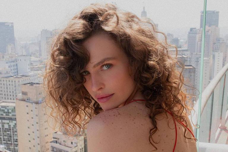 A imagem mostra uma mulher com cabelo cacheado e volumoso, usando um vestido vermelho com costas nuas. Ela está posicionada em uma varanda, olhando para a câmera, com um fundo de uma cidade grande, onde se podem ver prédios altos e uma atmosfera ensolarada