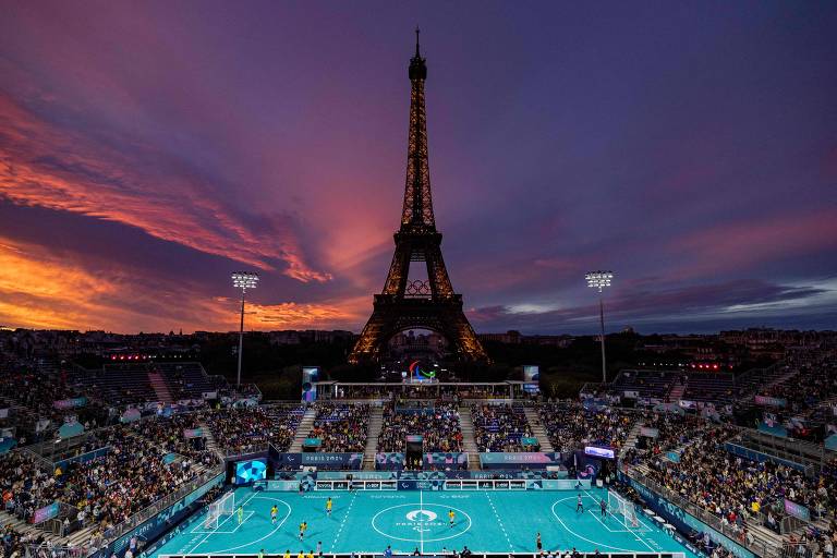 A imagem mostra um jogo de futebol em um campo com grama sintética azul, localizado sob a Torre Eiffel iluminada ao entardecer. O céu apresenta uma mistura de cores quentes, como laranja e roxo, enquanto uma grande quantidade de espectadores está presente nas arquibancadas. Há refletores iluminando o campo e uma tela de exibição visível. O evento parece ser parte das Olimpíadas de Paris 2024.
