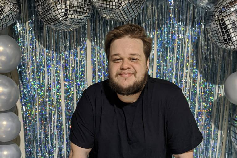 Um homem está sorrindo em frente a uma mesa com um bolo de aniversário prateado. O bolo tem uma decoração brilhante e uma vela com os números '28' e a frase 'Happy Birthday'. O fundo é decorado com balões prateados e uma cortina de papel metalizado que reflete a luz. O homem usa uma camiseta preta e está apoiado na mesa.