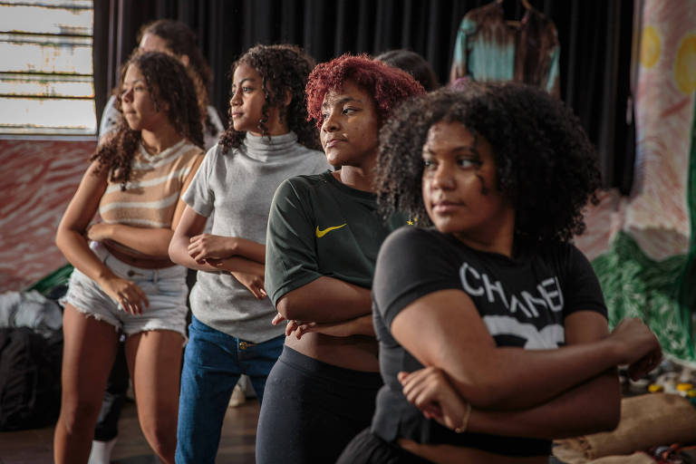 A imagem mostra um grupo de cinco jovens mulheres em um ambiente interno, possivelmente durante um ensaio. Elas estão alinhadas, com os braços cruzados e expressões concentradas. As mulheres têm cabelos cacheados e diferentes estilos de roupas, incluindo uma blusa cinza, uma camiseta preta com a palavra 'Chanel' e uma camiseta verde. Ao fundo, há uma parede decorada e alguns objetos no chão.
