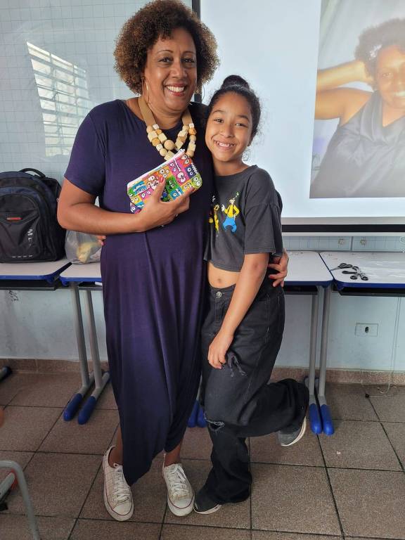 A imagem mostra uma mulher e uma menina sorrindo em um ambiente interno. A mulher, com cabelo cacheado e usando um vestido longo azul escuro, segura uma carteira colorida. A menina, com cabelo preso em um coque e vestindo uma camiseta preta, está ao lado da mulher, com um sorriso largo. Ao fundo, há uma tela com uma imagem de outra mulher. O ambiente parece ser uma sala de aula, com mesas e cadeiras visíveis.