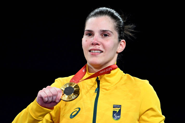 Uma atleta está sorrindo e segurando uma medalha de ouro. Ela usa uma jaqueta amarela com o logotipo do Brasil Paralímpico. A medalha tem um cordão vermelho com a inscrição 'PARIS 2024'. O fundo da imagem é escuro.
