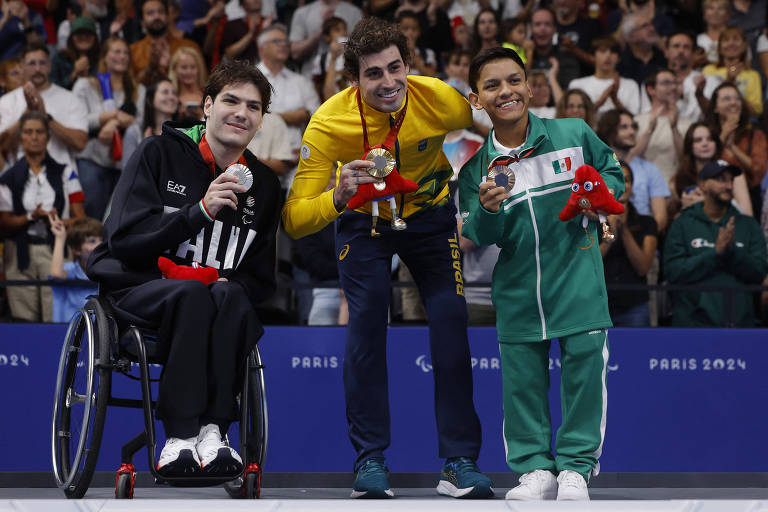 Três atletas estão no pódio, sorrindo e segurando suas medalhas. À esquerda, um atleta em cadeira de rodas, vestindo um moletom preto com a palavra 'ITALIA', segura uma medalha de prata. No centro, um atleta com uma camiseta amarela e calças azuis, representando o Brasil, exibe uma medalha de ouro. À direita, um atleta com um uniforme verde e branco, representando o México, segura uma medalha de prata e um brinquedo em forma de polvo. Ao fundo, uma plateia aplaude.
