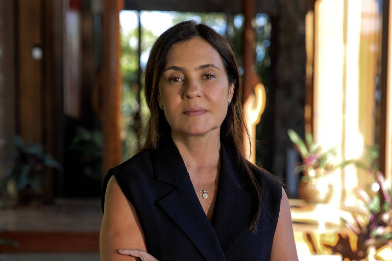 A imagem mostra uma mulher de pé em um ambiente interno, com paredes de madeira e grandes janelas que permitem a entrada de luz natural. Ela está usando um vestido preto sem mangas e tem os braços cruzados. Seu cabelo é longo e solto, e ela tem uma expressão séria.