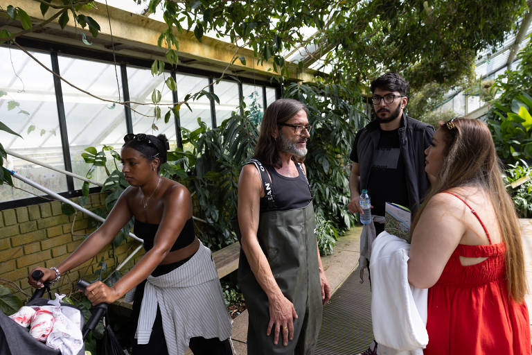 A imagem mostra um grupo de quatro pessoas em uma estufa com plantas. À esquerda, uma mulher de pele escura e cabelo preso está empurrando um carrinho. No centro, um homem de cabelo longo e barba está conversando com duas mulheres. Uma delas, à direita, tem cabelo longo e usa uma blusa vermelha, enquanto a outra, ao lado dela, usa uma blusa preta e tem cabelo castanho. O ambiente é verde e bem iluminado, com muitas plantas ao fundo.