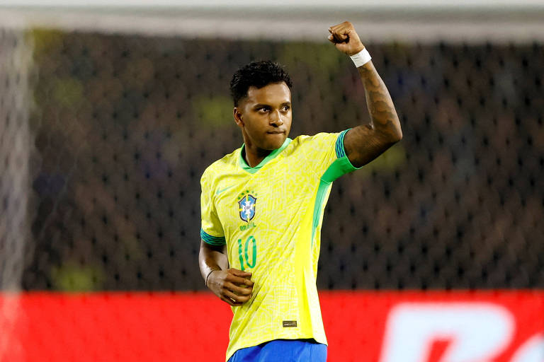 Um jogador de futebol está em campo, vestindo uma camisa amarela com detalhes em verde e azul, representando a seleção brasileira. Ele levanta o punho direito em sinal de celebração, com uma expressão de determinação no rosto. Ao fundo, há uma rede de gol e uma faixa vermelha com a palavra 'BRA' visível.
