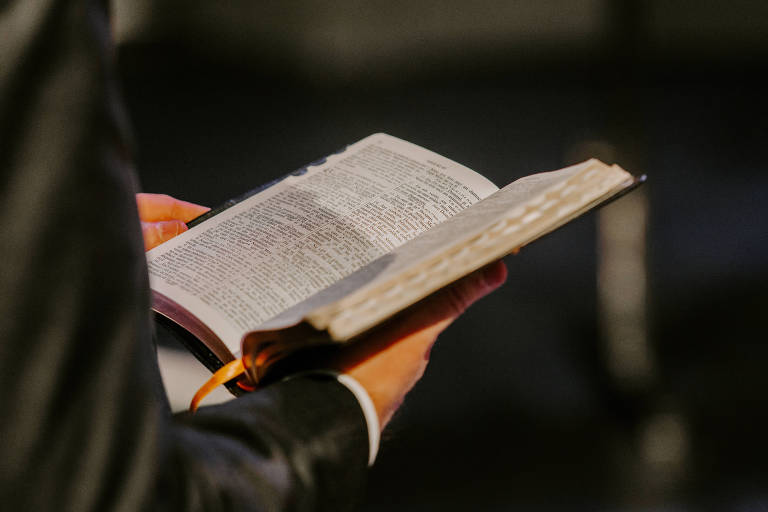 A imagem mostra uma pessoa de terno segurando uma Bíblia aberta com as duas mãos. A Bíblia está em foco, enquanto o fundo é desfocado, sugerindo um ambiente de culto ou reunião. A mão esquerda da pessoa está na parte superior da Bíblia, enquanto a mão direita segura a parte inferior