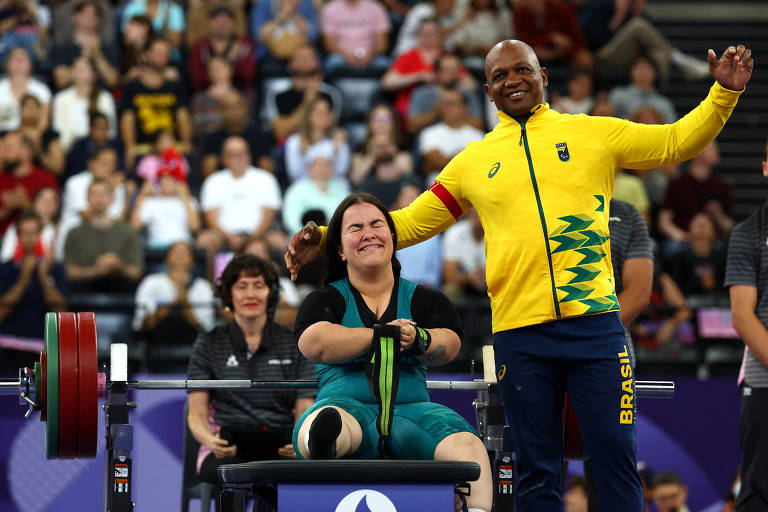 A imagem mostra uma atleta de levantamento de peso sentada em um banco, sorrindo e segurando um peso com as mãos. Ela está vestindo um uniforme azul e preto. Ao lado dela, um treinador, vestido com uma camisa amarela e azul com a palavra 'BRASIL', está levantando a mão em celebração. Ao fundo, uma plateia assistindo ao evento.
