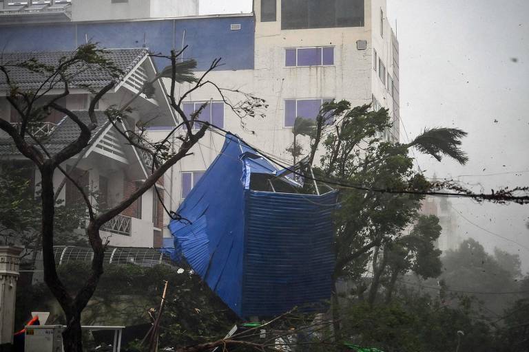 A imagem mostra uma cena de destruição causada por um evento climático severo. Um grande pedaço de material azul, possivelmente uma cobertura ou lona, está preso em uma árvore, enquanto ao fundo se vê um edifício com janelas quebradas. Árvores estão inclinadas devido ao vento forte, e o céu está nublado, indicando condições climáticas adversas.