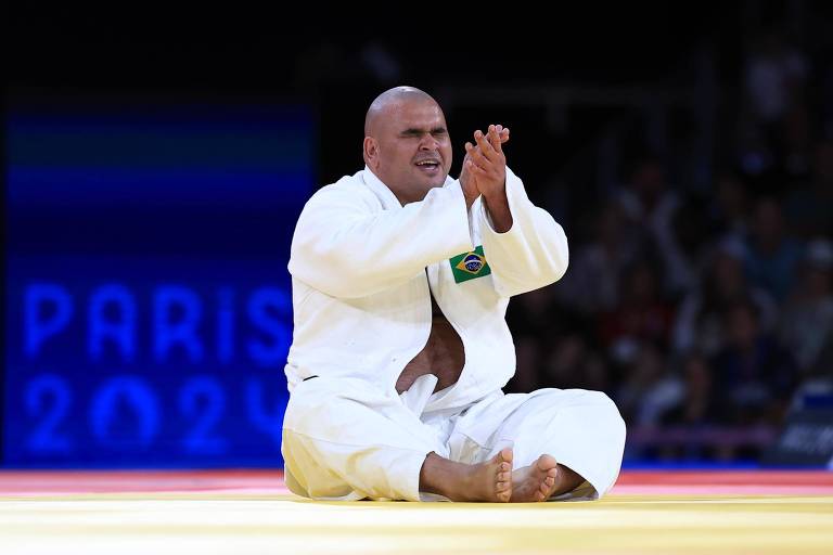 Um atleta de judô está sentado no tatame, aplaudindo. Ele usa um judogi branco e tem uma bandeira do Brasil em sua roupa. O fundo mostra uma tela com as palavras 'PARIS 2024'. O tatame é colorido, com áreas em vermelho, amarelo e azul.