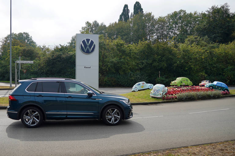 A imagem mostra um carro SUV da Volkswagen, estacionado em uma estrada, com um grande logotipo da Volkswagen ao fundo. À direita, há uma área ajardinada com várias tendas coloridas dispostas em um círculo. O ambiente é predominantemente verde, com árvores ao fundo e um céu nublado.