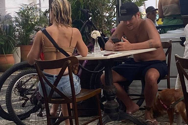 A imagem mostra um café ao ar livre com duas pessoas sentadas em uma mesa. Uma mulher de costas, com cabelo molhado e usando um biquíni, está sentada à esquerda. Um homem, sem camisa e usando um boné, está à direita, olhando para o celular. Há uma flor em um vaso pequeno sobre a mesa. Ao fundo, pode-se ver uma bicicleta encostada e outras pessoas, além de palmeiras e uma estrutura de toldo.