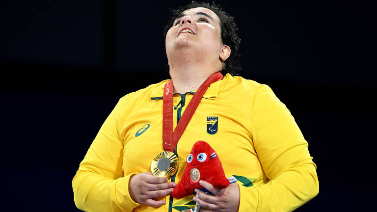 Uma atleta vestindo uma camisa amarela com detalhes verdes, segurando uma medalha dourada e um boneco vermelho. Ela está olhando para cima, com uma expressão de alegria e emoção. A medalha está pendurada em seu pescoço por uma fita vermelha.
