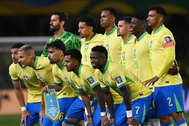 A imagem mostra a equipe de futebol do Brasil posando para uma foto. Os jogadores estão em duas fileiras, com os da frente agachados e os de trás em pé. Todos estão vestindo uniformes amarelos com detalhes verdes e azuis. Um dos jogadores segura uma bandeira com o emblema da CBF e informações sobre a Copa do Mundo. O goleiro está atrás, usando um uniforme verde.
