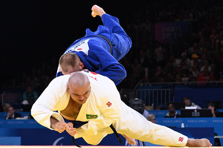 A imagem mostra dois lutadores de judô em ação. Um lutador, vestido com um judogi branco e com a bandeira do Brasil, está se abaixando enquanto o outro, vestido com um judogi azul, está saltando sobre ele. O fundo é uma arena com espectadores e equipamentos de filmagem visíveis.
