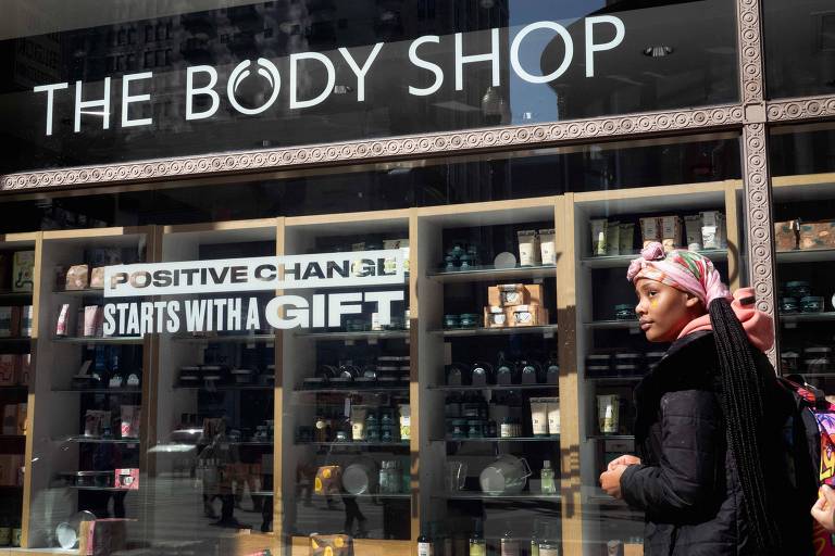 A imagem mostra a vitrine da loja The Body Shop, com prateleiras exibindo produtos de beleza. Um cartaz na vitrine diz 'POSITIVE CHANGE STARTS WITH A GIFT'. Uma mulher com um lenço na cabeça está parada ao lado da vitrine, olhando para os produtos.