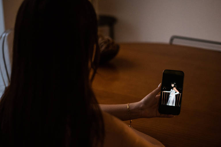 A imagem mostra Camila de costas, com cabelo longo e escuro, olhando para a tela de um celular. Na tela do celular, é visível uma imagem de de uma garota vestida de branco para uma festa de debutante. É Camila mais jovem. O ambiente é interno, com uma mesa de madeira ao fundo e uma iluminação suave.