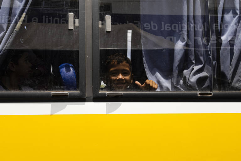 A imagem mostra uma criança sorrindo e fazendo um sinal de positivo através da janela de um ônibus. A parte inferior do ônibus é amarela, e as cortinas da janela estão parcialmente abertas, permitindo a visão do interior. Ao fundo, é possível ver outras pessoas dentro do ônibus