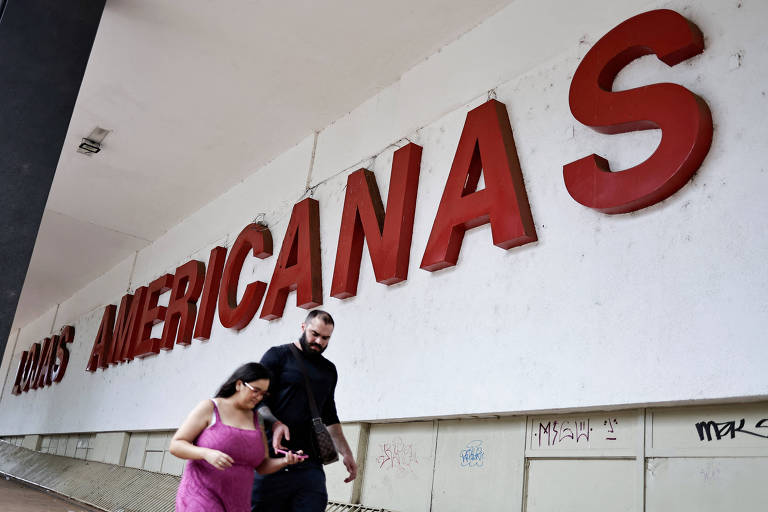 A imagem mostra a fachada de uma loja chamada 'Lojas Americanas', com letras grandes e vermelhas. Na parte inferior, duas pessoas estão caminhando, uma mulher vestindo uma blusa rosa e um homem ao seu lado. O fundo é uma parede branca com algumas marcas e grafites.